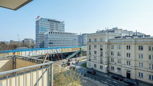 A multi-story building with a modern glass facade, a pedestrian overpass, and a street with parked cars. - RentPlanet - Studio Obozowa (Warsaw)