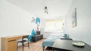 A room with a wooden desk, a bed with a blue bedspread, a dining table with a green plate, and a window with blinds. - RentPlanet - Studio Obozowa (Warsaw)