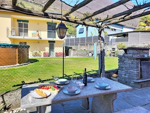Ein Essbereich im Freien mit einem Steintisch, umgeben von einem Maschendrahtzaun, mit Blick auf ein gelbes Gebäude und eine Steinmauer im Hintergrund.