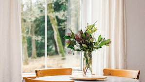 Une table à manger ornée d’un vase de fleurs, deux chaises, et une vue sur les arbres par la fenêtre.