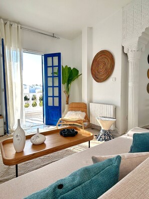 A living room with a blue door, a wicker chair, a small table, and a sofa with teal cushions. - Aurea Apartment in the Heart of Sidi Bou Saïd (La Marsa)