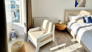 Ett modernt sovrum med en säng, en stol, ett litet bord, en vas och en tavla.