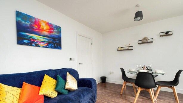 A modern living room with a blue sofa, colorful cushions, a glass dining table with chairs, and a painting on the wall. - Freya`s Flat with Free Parking (Leicester)