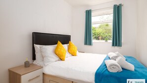 A hotel room with a bed, a nightstand, a window with curtains, and a view of greenery outside. - Freya`s Flat with Free Parking (Leicester)