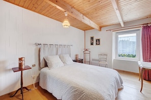 Un dormitorio con techo de madera, cama con colcha estampada, mesita de noche con lámpara y ventana con cortinas.