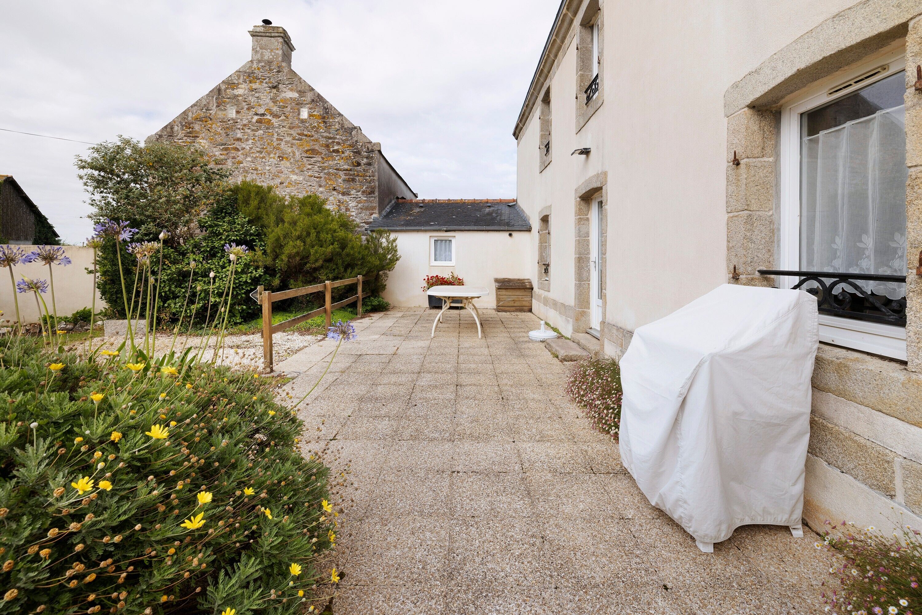 Un bâtiment en pierre doté d’un patio carrelé, d’une table couverte et d’une fenêtre avec un rideau.