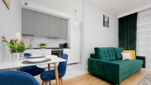 Un salon moderne avec un canapé vert, des fauteuils bleus, une table à manger dressée pour deux personnes et une cuisine en arrière-plan.