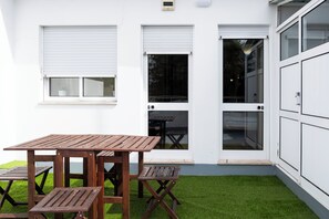 Eine Terrasse mit Holztischen und -stühlen, einem kleinen weißen Tisch und einer Tür zu einem überdachten Bereich.