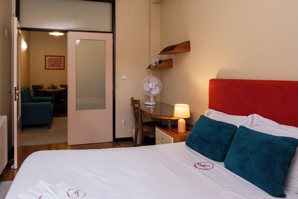 Una habitación de hotel con una cama, un escritorio con una silla, una lámpara y un ventilador.