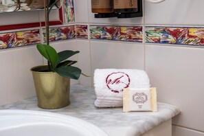 Ein Badezimmer mit Marmorwaschbecken, einer Topfpflanze, einem weißen Handtuch mit rotem Logo und einer Seifensäckchen.