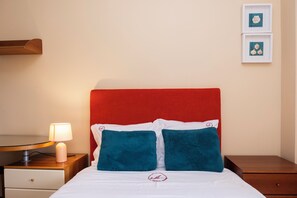 Un lit bien fait, avec une tête de lit rouge, une literie blanche et deux coussins turquoise. On aperçoit une lampe de chevet à gauche, une petite table de chevet en bois et un tableau encadré au mur.