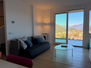 Ein modernes Wohnzimmer mit einer Couch, einem kleinen Tisch und Blick auf eine gebirgige Landschaft durch große Fenster.
