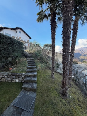 Eine steinerne Treppe führt zu einem Haus mit Balkon, umgeben von Grünpflanzen und Palmen.