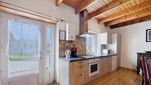 A kitchen with wooden cabinets, a built-in oven, a coffee maker, and a window with a view of a garden. - Ty izella – 500m from the sea (Plomodiern)