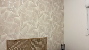 Ein Schlafzimmer mit Wand im geometrischen Muster, einem Holzkopfende, einem Bett mit beige farbenem Bettbezug und einem Nachttisch mit Lampe.