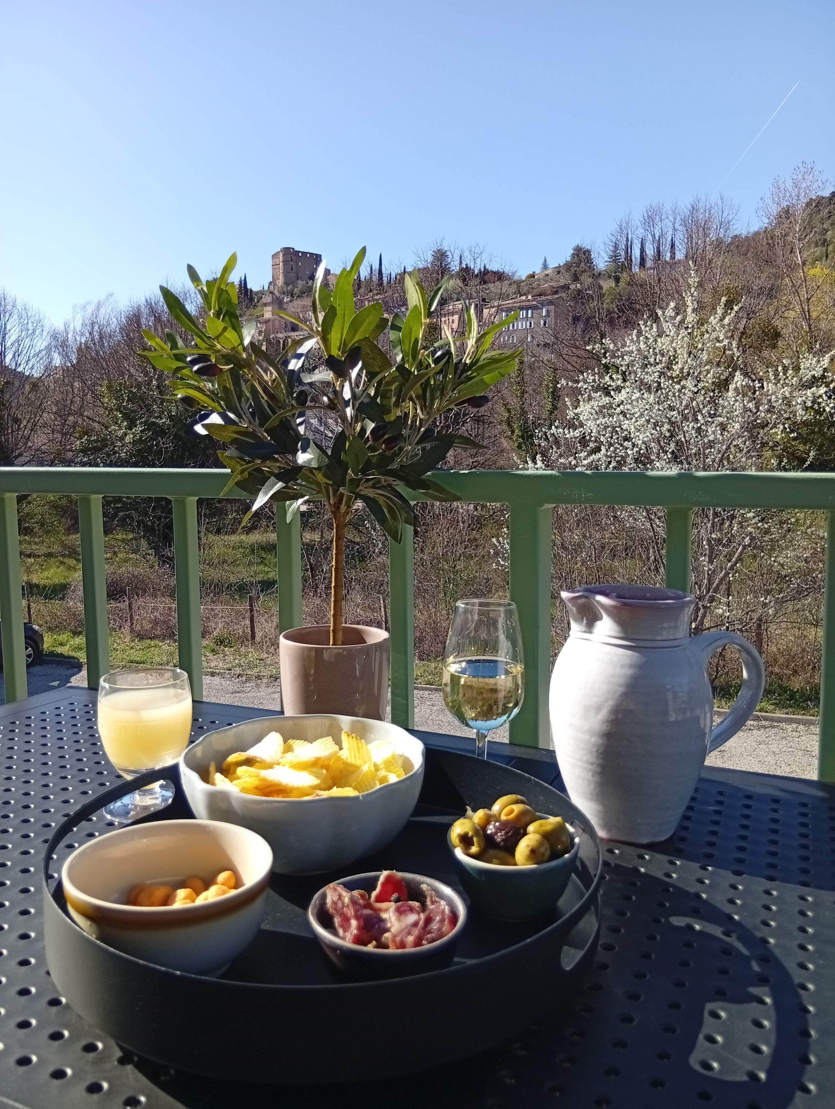 A table with food and drinks, a potted plant, and a view of a hill with buildings.