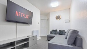 Ein Zimmer mit einem Flachbildfernseher, der 'NETFLIX' zeigt, einem grauen Sofa mit Kissen, einem kleinen Tisch und einer weißen Tür.