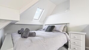 Ein Schlafzimmer mit schrägen Wänden, einem Dachfenster, einem Bett mit zusammengerollten Handtüchern, einem Nachttisch und einer Schubladenkommode.