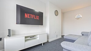 Ein modernes Wohnzimmer mit einem Wand-Fernseher, einer weißen Unterhaltungseinheit, einem grauen Sofa, einer Uhr und einem Schminkspiegel.