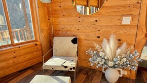 Interior acollidor d'una cabana amb parets de fusta, un mirall rodó, una petita cadira, una catifa de pell i un vas amb plantes seques.