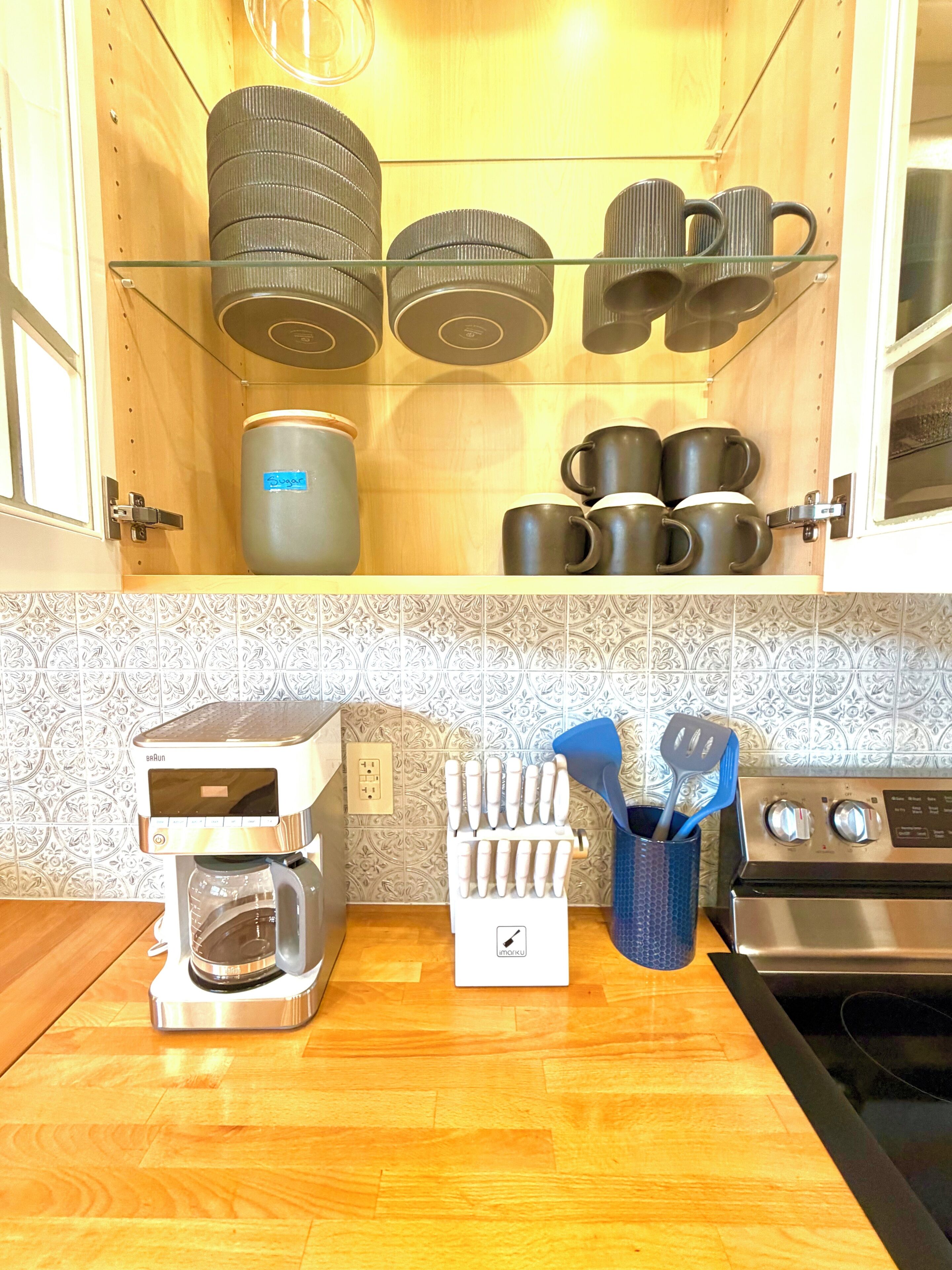 Cozinha com cafeteira, porta-utensílios e conjunto de canecas nas prateleiras.