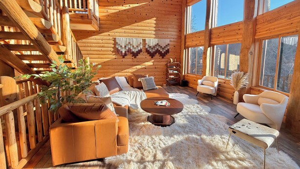 Interior d'una cabana de fusta amb una escala de fusta, seients de sofà, una taula de centre i una finestra gran.