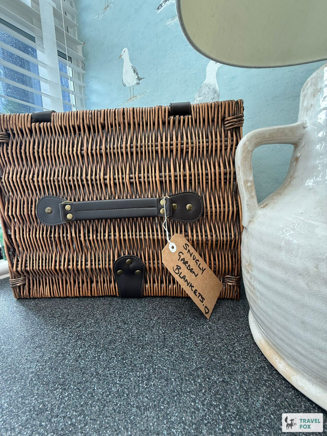 Une valise en osier avec une étiquette indiquant « Couvertures confortables pour le jardin » et un pichet en céramique blanche.