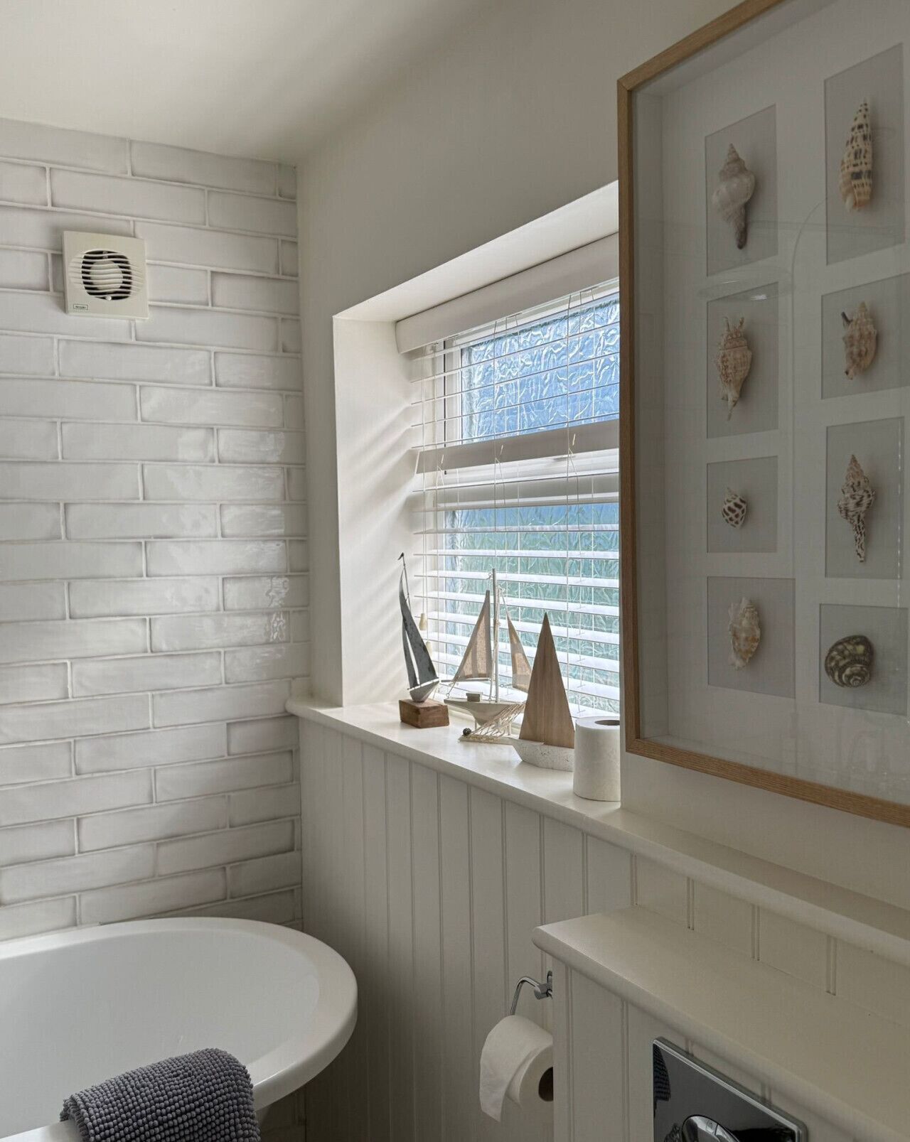 Un baño con pared de ladrillo blanco, una ventana con vistas, un lavamanos y una colección enmarcada de caracolas.