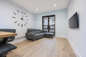 A modern living room with a grey sectional sofa, a wooden dining table, a large wall clock, and a TV mounted on the wall. - Matilda`s Central Stay (Nottingham)