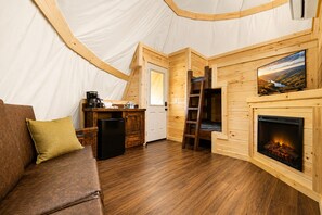 A tented accommodation with a wooden interior, a sofa, a kitchen area, a fireplace, and a ladder.