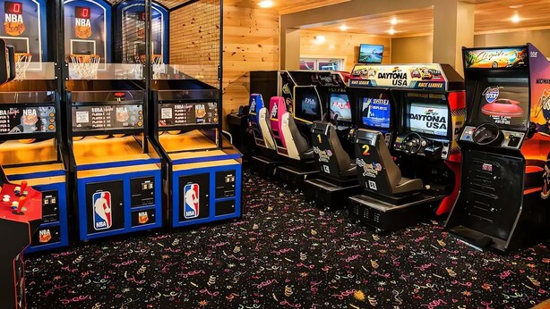 Eine Reihe von Spielautomaten, darunter NBA und Daytona USA, mit einer Holzbalkendecke und einem gemusterten Teppich.