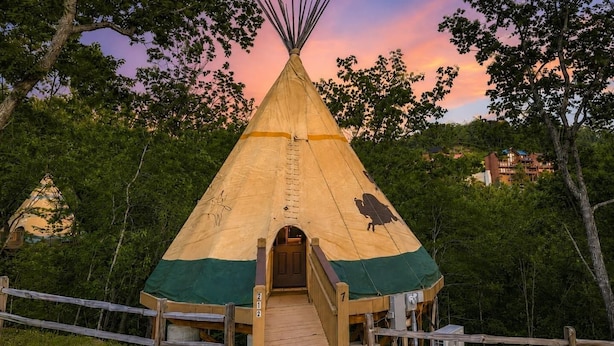 A teepee with a wooden deck leading to the entrance, surrounded by trees and a building in the background.