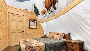 Ein Zelt mit Holzrahmen, ausgestattet mit einem Bett, einem Nachttisch und einer Wand mit einer Kuh- und Baummalerei.