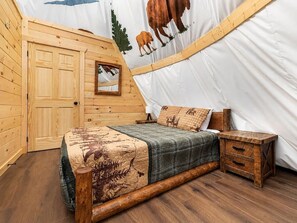 A wooden-framed tent with a bed, a nightstand, and a wall with a cow and tree mural. - Unique Glamping + Resort Fun access •Sleep 4 (Sevierville)