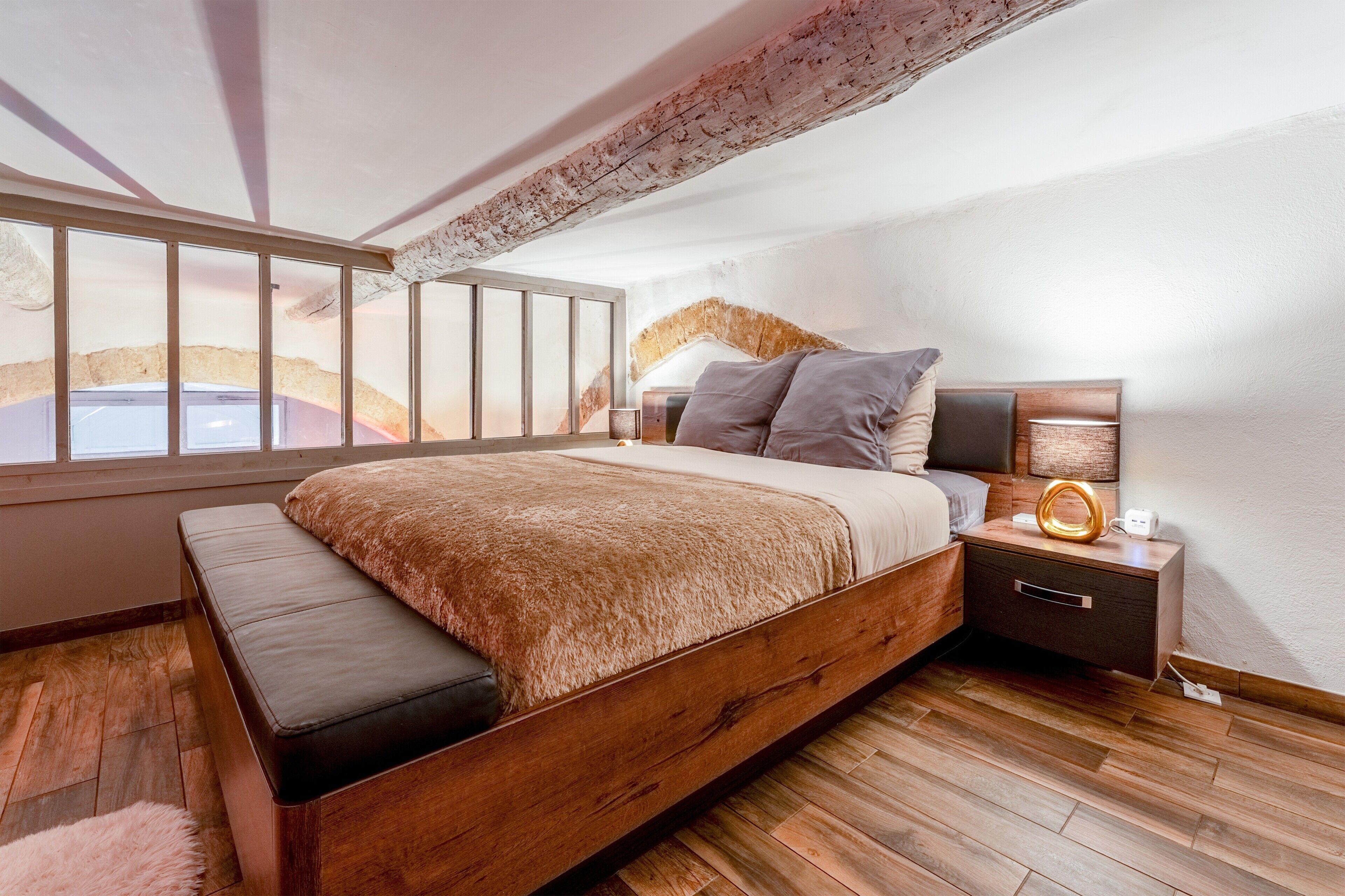 A bedroom with a wooden bed, a bench, a nightstand with a lamp, and a view of the outdoors through large windows.