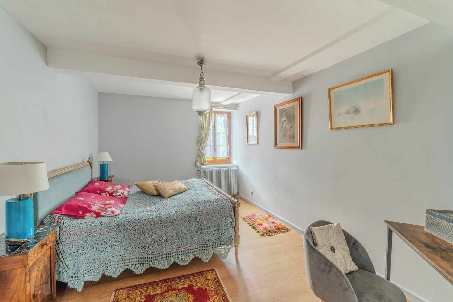 Un dormitorio con cama, mesita de noche, lámpara, cómoda, silla, ventana con cortinas y radiador.