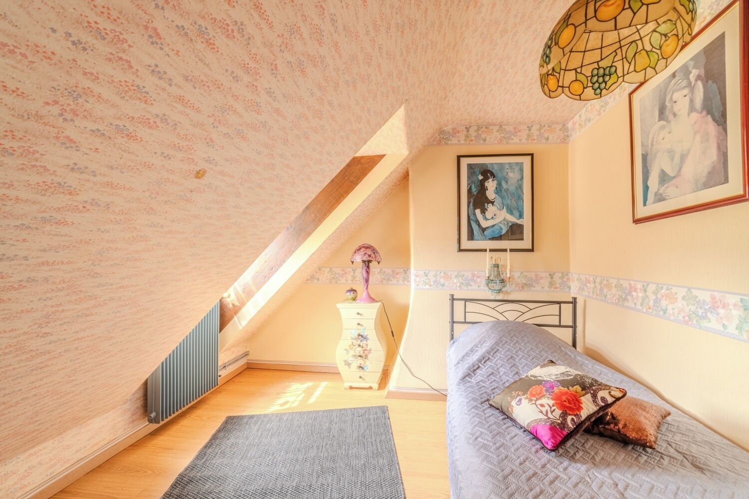 Una camera da letto con soffitto spiovente, un letto con cuscini floreali, una cassettiera, un quadro e una lampada con vetrate artistiche.