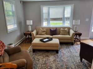 Un salon avec un canapé beige, un pouf, un tapis à motifs, une chaise en bois et une fenêtre avec des stores.