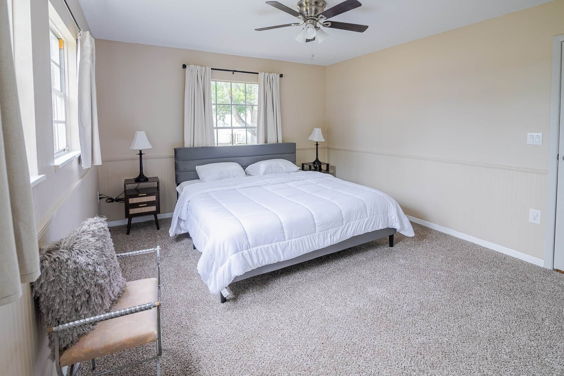 Une chambre à coucher comprenant un lit, deux tables de chevet, une chaise, un ventilateur de plafond et une fenêtre avec des rideaux.