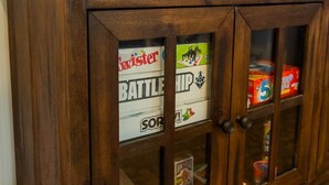 Un meuble en bois doté de portes en verre, contenant divers objets, comme un jeu de Twister, un jeu de Bataille navale et un paquet de chewing-gum à 5 cents.