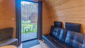 A cozy cabin interior with a wooden ceiling, a black leather sofa, a small table, and a view of an outdoor area with a picnic table and a wooden structure. - Zeeland Glamping Lodge for 2 (Burgh-Haamstede)