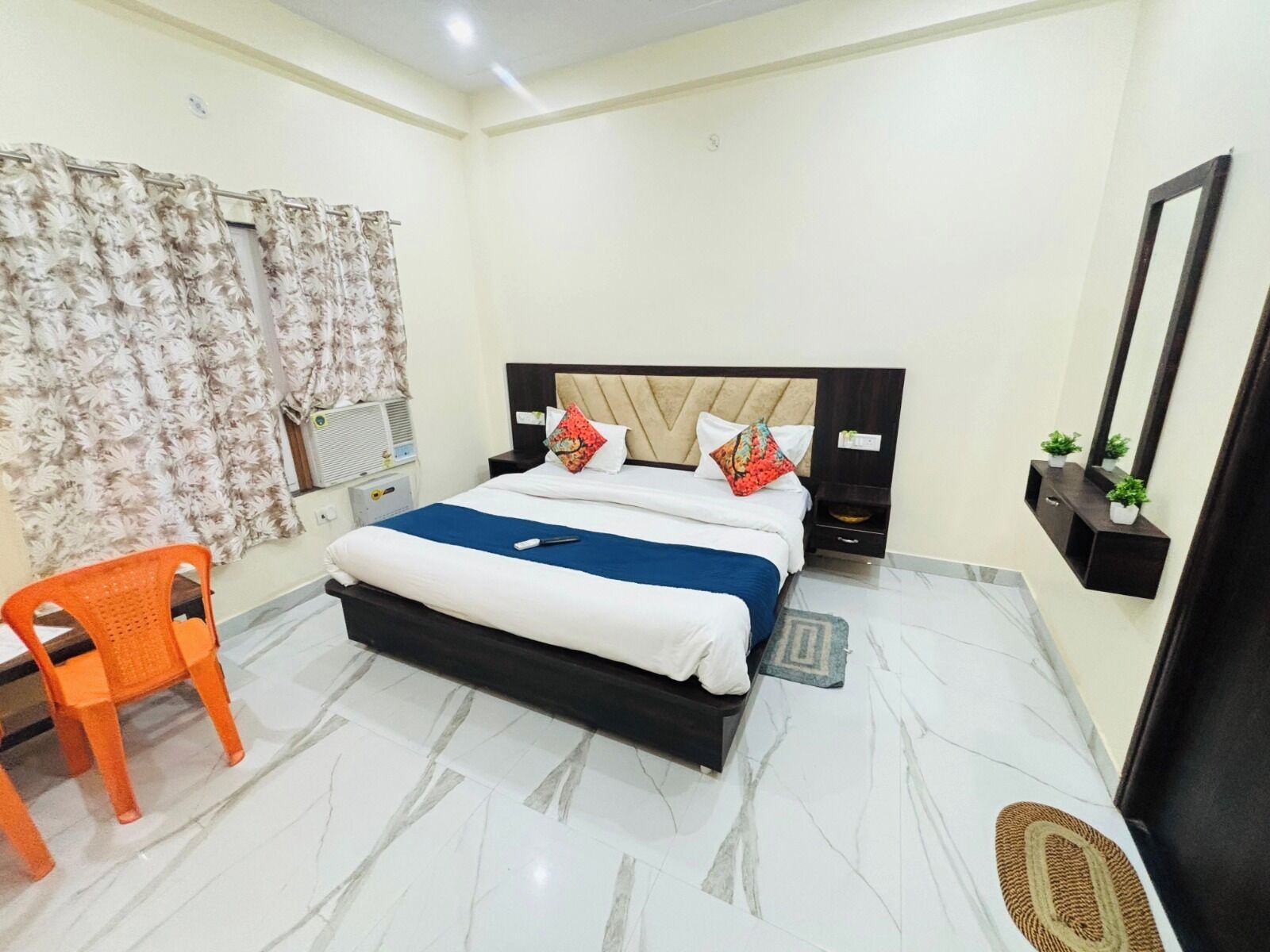 Habitación de hotel con cama, una silla, una mesita de noche, un espejo y un estante en la pared con plantas.