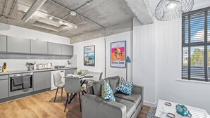 A modern kitchen with grey cabinets, a dining area with a grey sofa and table, and a window with blinds. - Apartment 18 - The Works (Woolston)