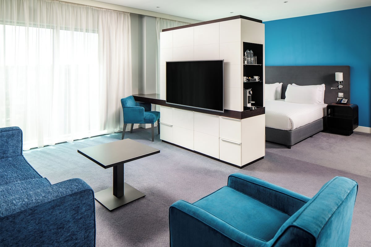 A modern hotel room with a flat-screen TV, a bed, a desk, and two blue armchairs.