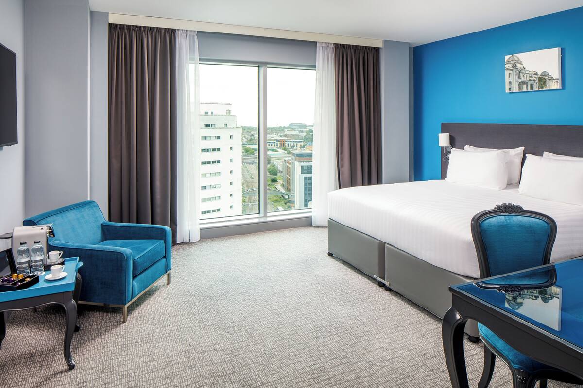 A hotel room with a large bed, a blue armchair, a small table with a coffee maker, and a view of buildings through the window.