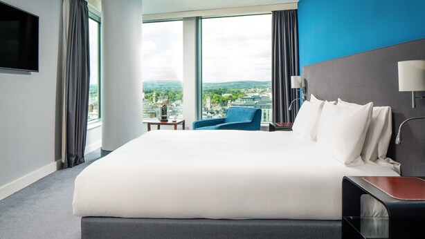 A hotel room with a large bed, a blue chair, a TV, and a city view. - Majestic Hotel Cardiff (Cardiff)