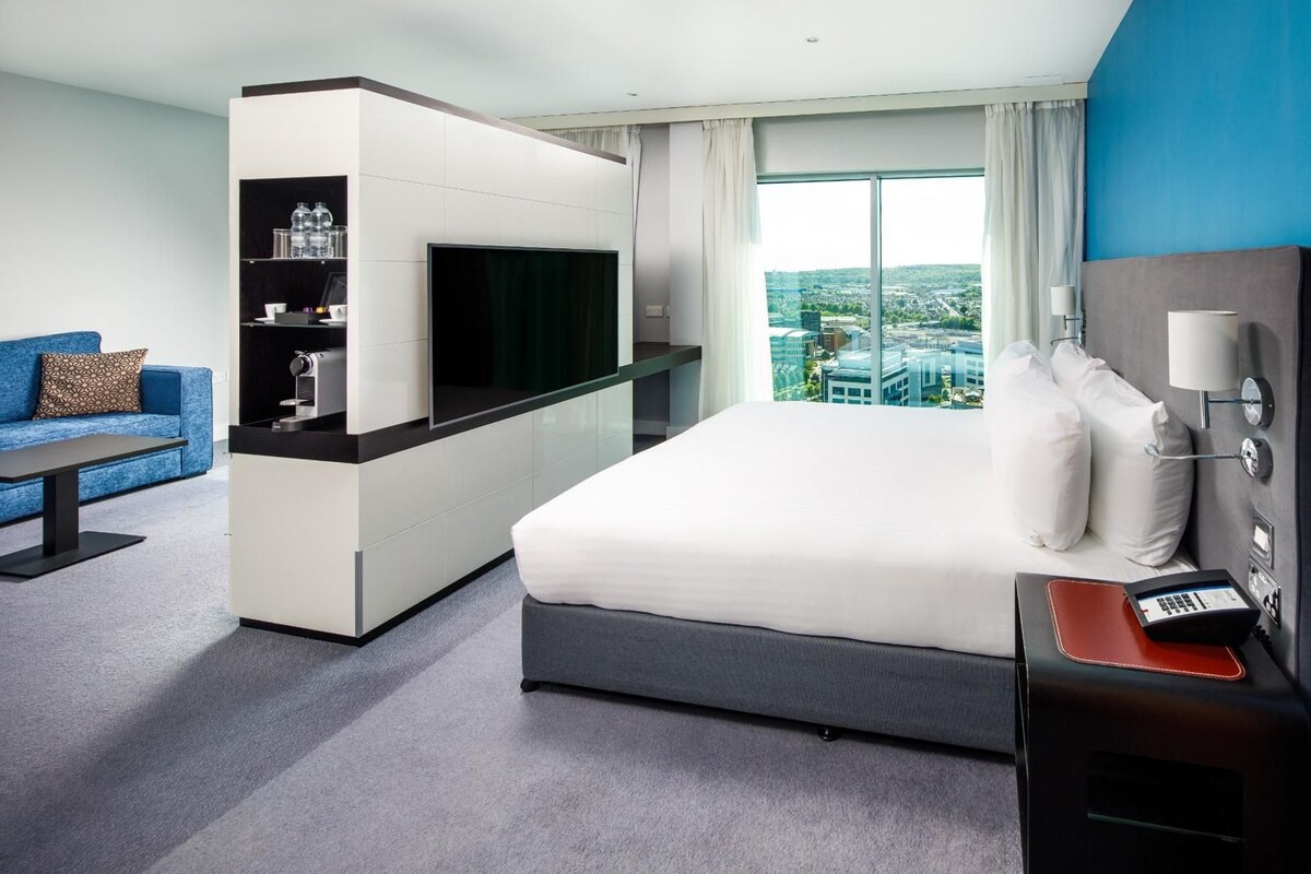 A hotel room with a bed, a TV, a desk, a sofa, and a view of a cityscape.