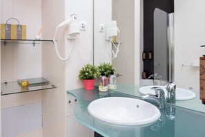 Un baño moderno con dos lavamanos, una encimera de vidrio, una planta en maceta y secadores de pelo montados en la pared.