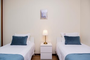 A hotel room with two beds, a nightstand with a lamp, a small framed picture on the wall, and a white door.