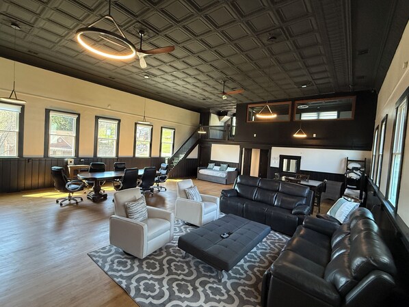 A spacious living area with a high ceiling, large windows, a seating arrangement, and a staircase leading to an upper level.